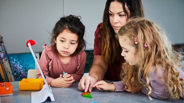 En pedagog och två barn tittar på en skärm och pekar på former i olika färger som ligger på bordet.