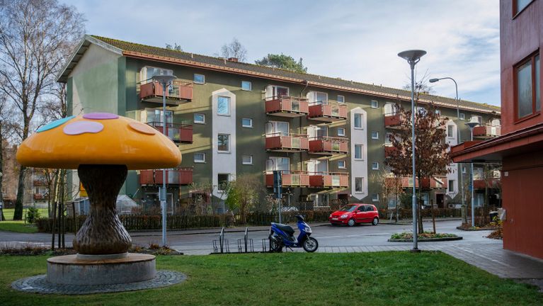 Ett bostadsområde där 50-tals hus syns bakom en stor lekfull svamp med gul hatt och prickar i pastellfärger.