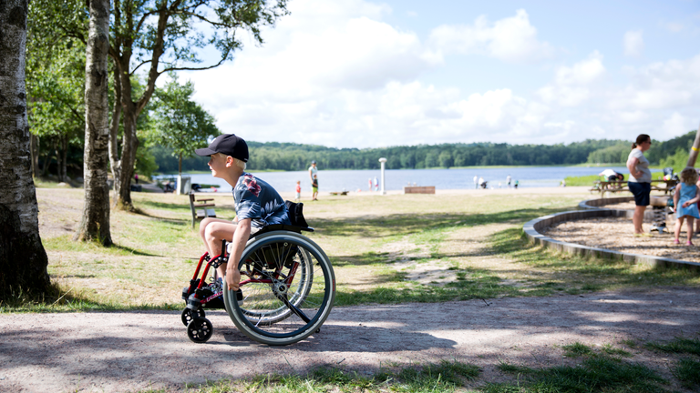 En pojke i rullstol på en stig, med en badplats i bakgrunden.